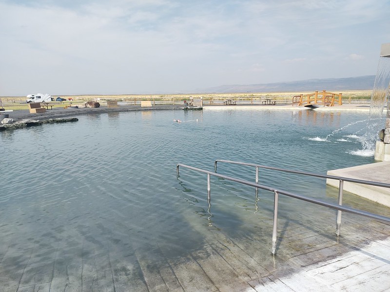 A large pool, surrounded by cement, with a desert landscape in the background. A tiny image of a woman is in the middle.